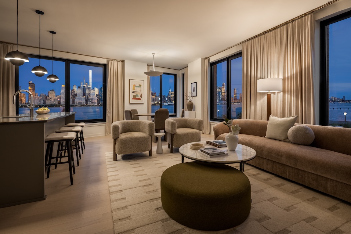 Open-concept living room and kitchen with floor-to-ceiling windows overlooking a waterfront New York City skyline.