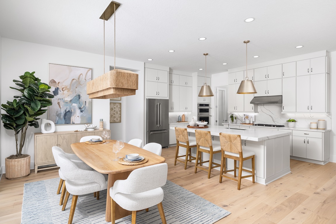 Breakfast nook with a light wood dining table, curved upholstered chairs, woven pendant light, and an adjacent white kitchen.