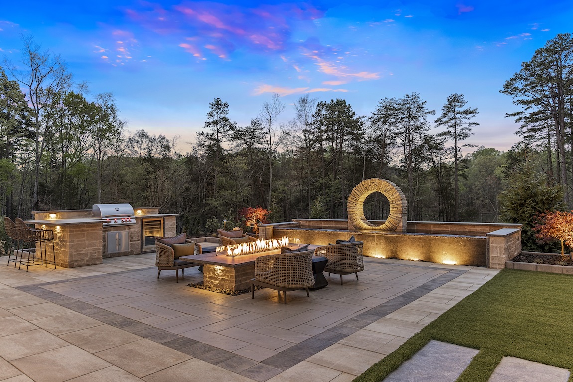 Luxury backyard central fire pit seating, grilling station, unique water feature, and landscaped grounds.