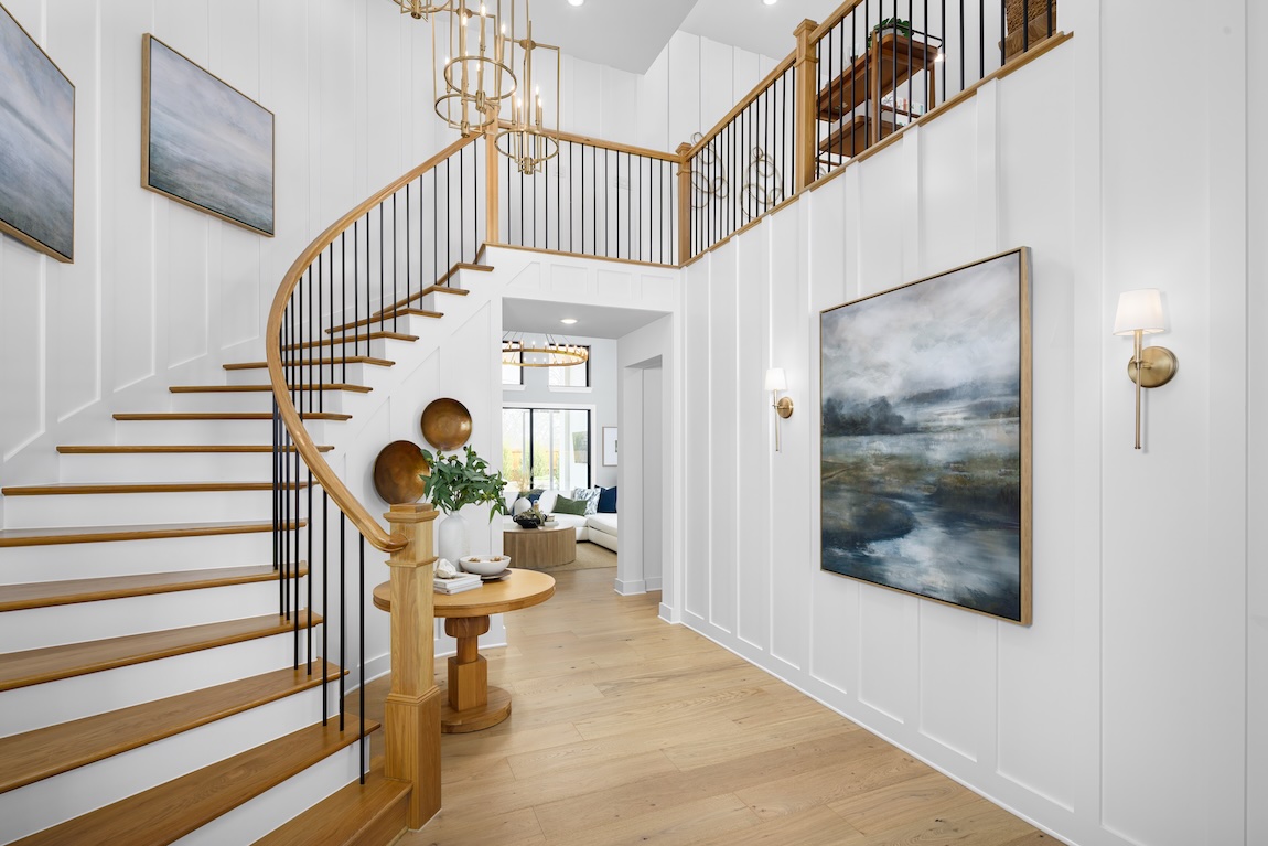 Curved wooden staircase with black spindles in a white paneled foyer featuring brass lighting, landscape artwork, and warm natural flooring