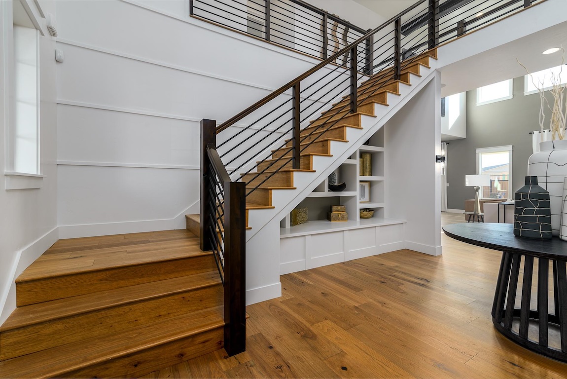 Modern foyer with a wood staircase featuring horizontal metal railings, built-in shelving beneath the stairs, white paneled walls, and warm hardwood flooring.