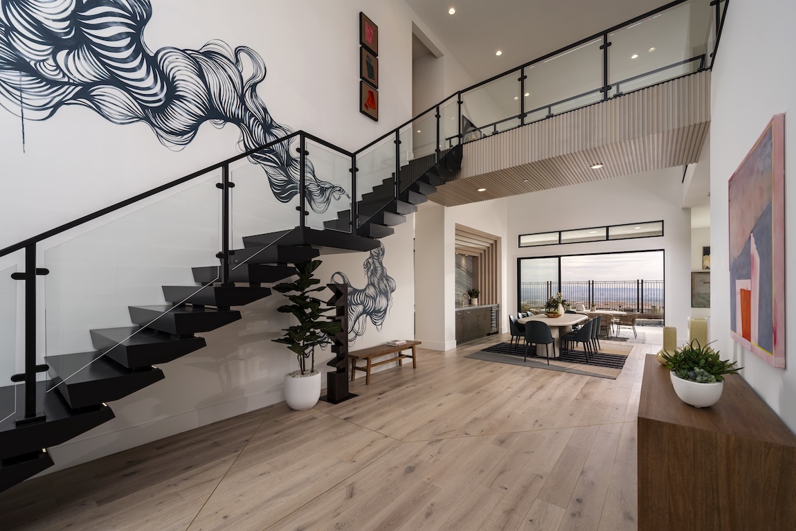 Modern entryway with an open black floating staircase, glass railings, bold abstract wall mural, light wood flooring, and a dining area visible beyond.