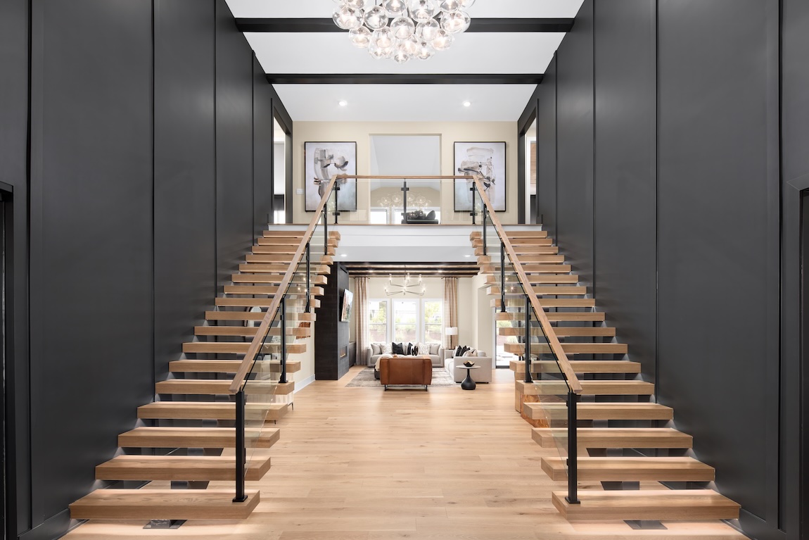 Grand two-story foyer with a floating double staircase, light wood treads, glass railings, dark accent walls, and a modern chandelier opening to a living area beyond.