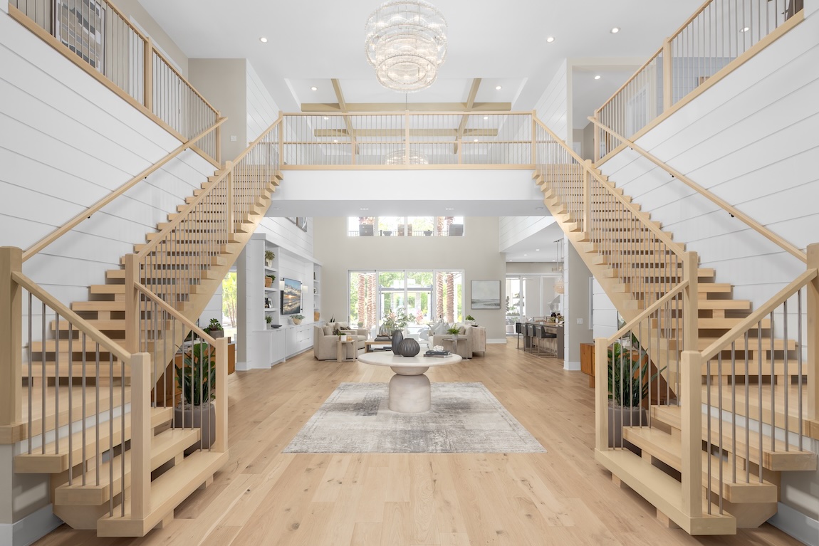 Grand double staircase in light wood framing a modern open-concept living room with vaulted ceiling and chandelier centerpiece