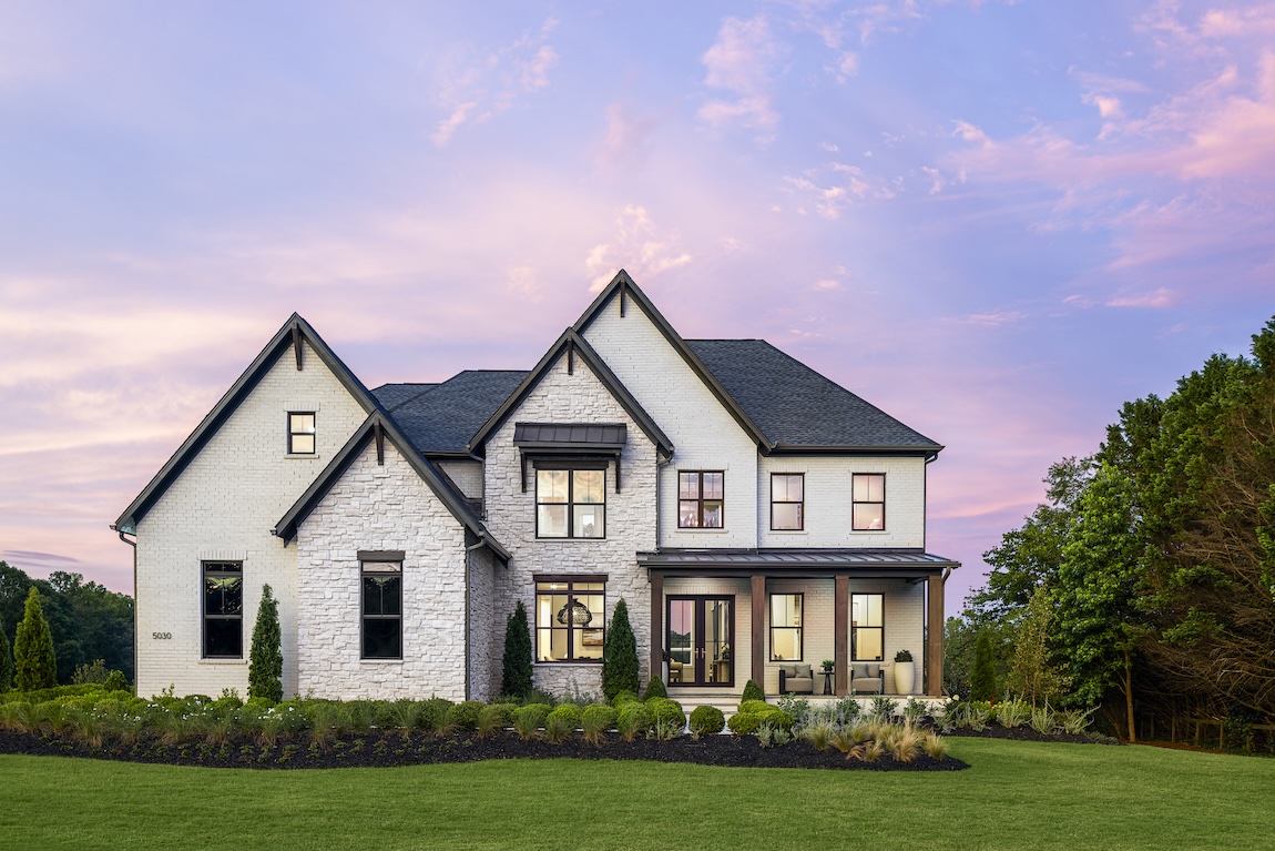 Luxury farmhouse-style home exterior with white brick, stone accents, covered front porch, and landscaped lawn at sunset.