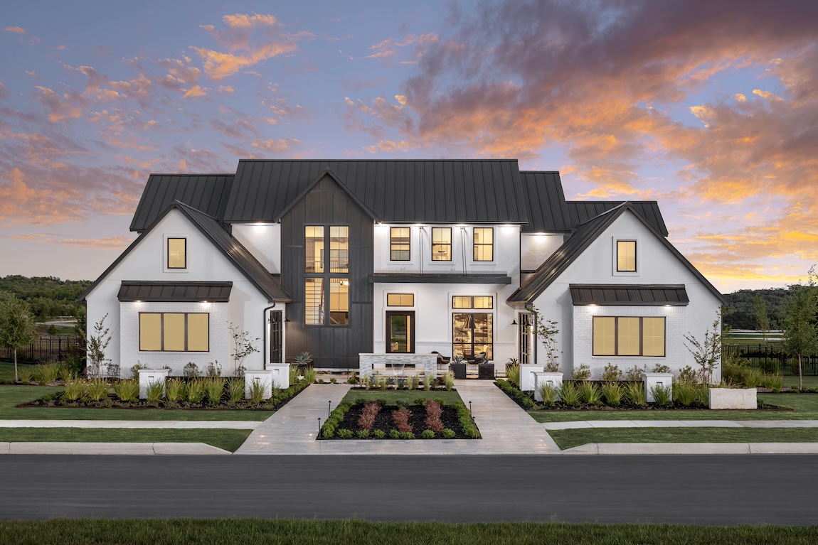 Modern farmhouse exterior with black metal roof and white brick facade, lit at twilight with symmetrical landscaping and sunset sky backdrop
