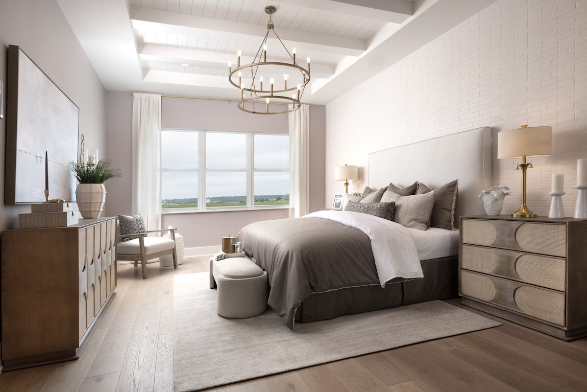 Serene primary bedroom with soft neutral tones, natural wood textures, vaulted ceiling with chandelier, and a wide window overlooking scenic landscape in a Toll Brothers home.