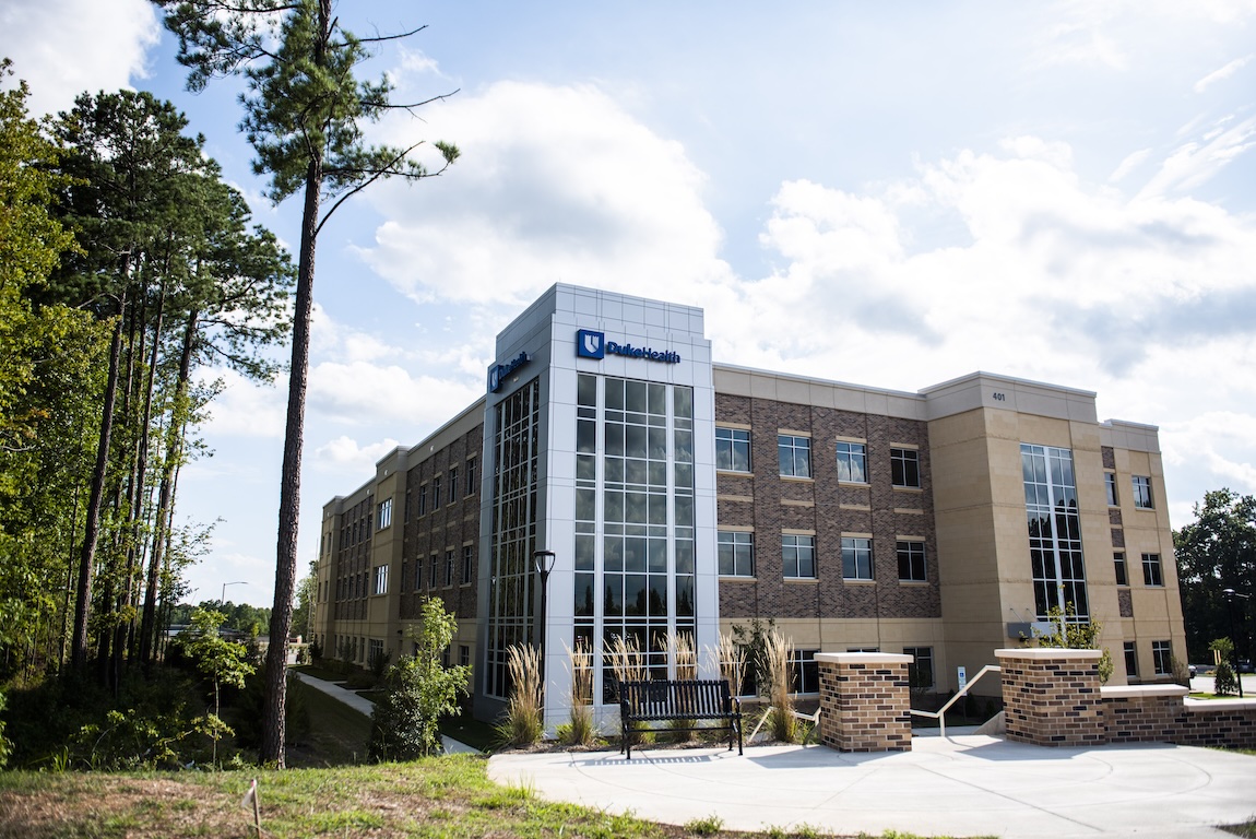 Modern medical office building with a glass entry tower, brick and stone exterior, landscaped walkways, and tall pine trees under a partly cloudy sky.