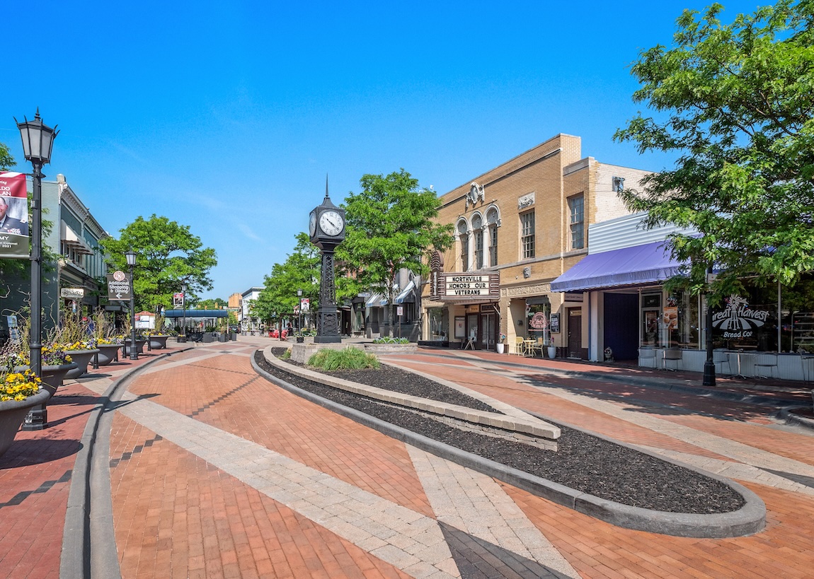 Downtown streetscape with brick-paved walkways, a central clock tower, tree-lined sidewalks, and historic storefronts under a clear blue sky.