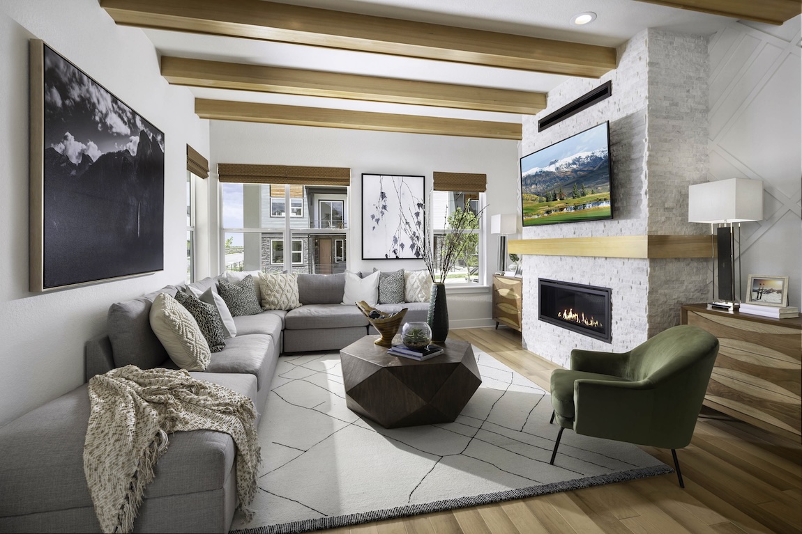 Cozy great room with exposed wood ceiling beams, a stone fireplace with mounted TV, sectional seating, accent chairs, and large windows bringing in natural light.