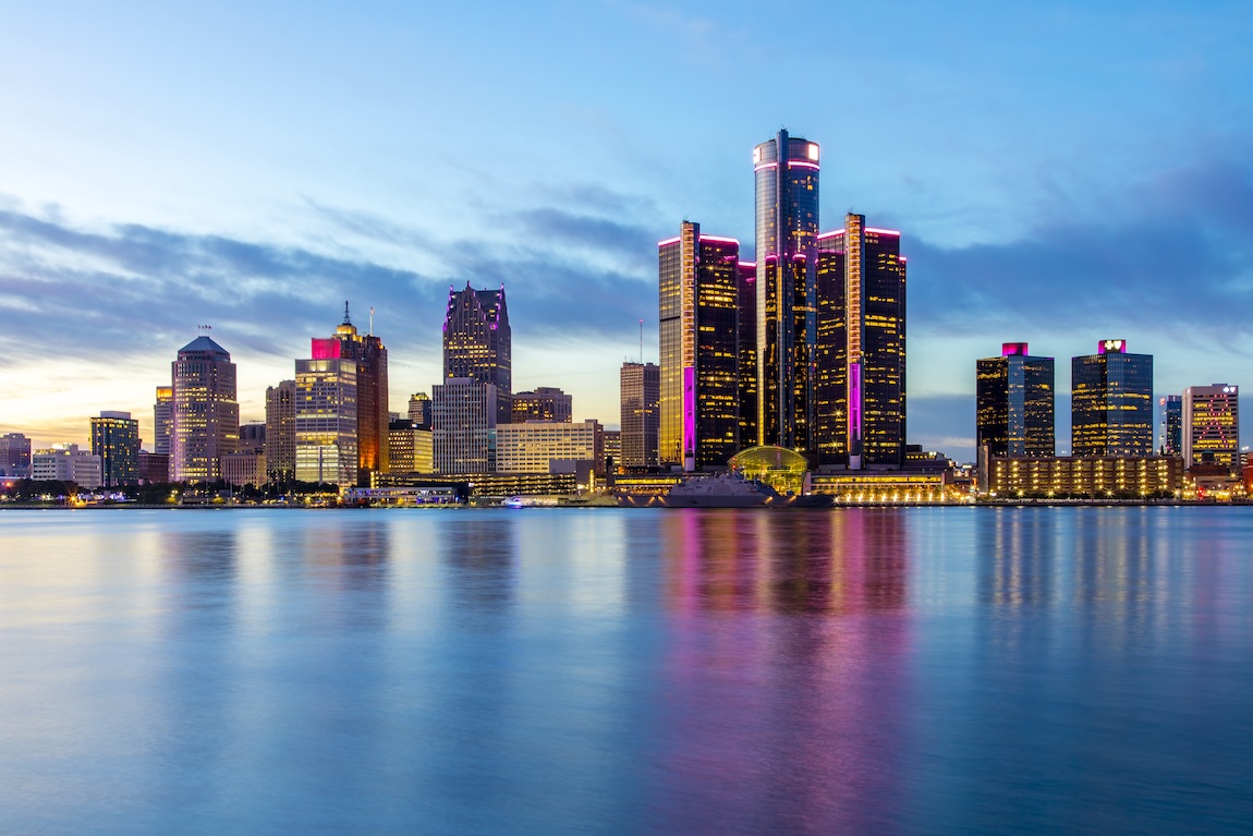 The skyline of Detroit in the evening, with pink lights on many of the buildings for breast cancer awareness. The skyline is reflecting off the Detroit River.