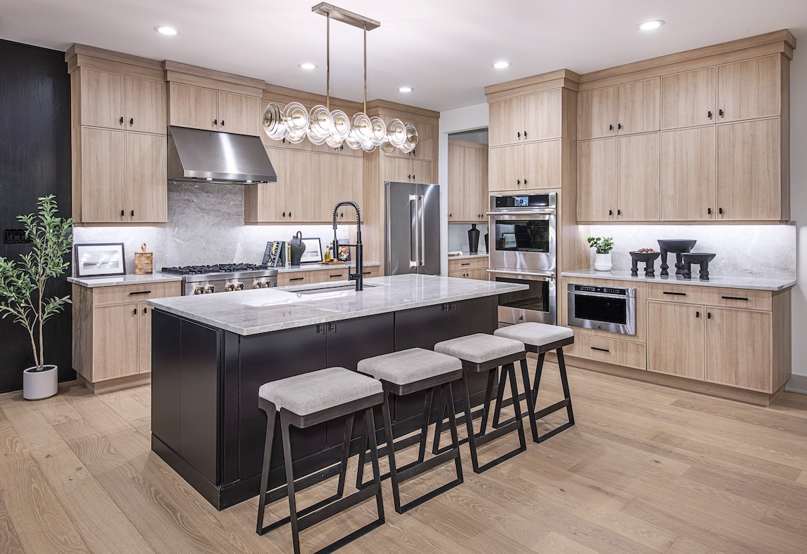 Modern kitchen with light wood cabinetry, a contrasting dark island with seating, stainless steel appliances, statement linear pendant lighting, and hardwood floors.