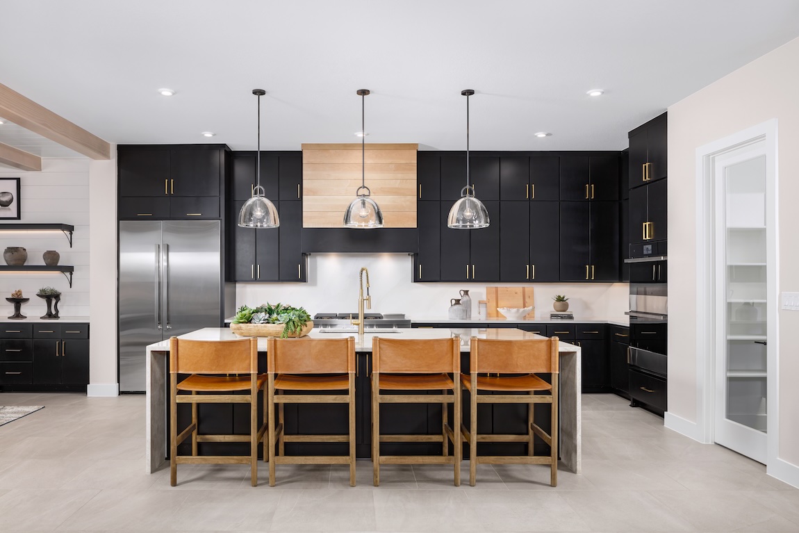 Spacious modern kitchen with black cabinetry, brass hardware, a white island with leather bar stools, wood-paneled range hood detail, and open shelving accents.