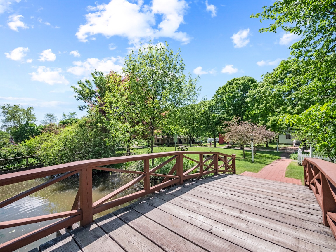 Wooden pedestrian bridge spanning a calm river in a green park setting, surrounded by mature trees, walking paths, and spring foliage.