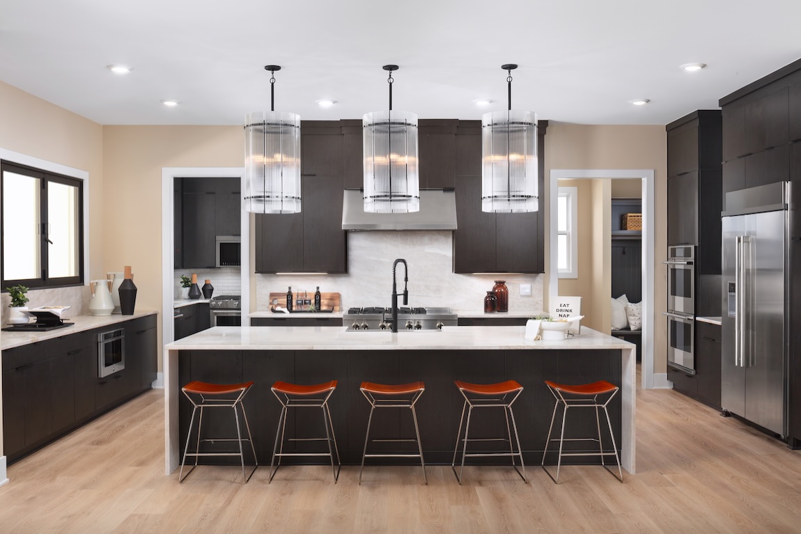 Sleek kitchen design with a wide white island, dark wood cabinetry, cylindrical glass pendant lights, stainless steel appliances, and light wood flooring.