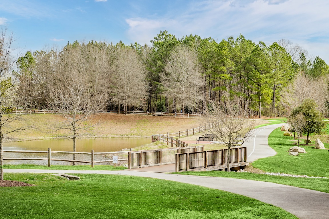 Paved walking trail curving through a park with a wooden footbridge, pond, open lawn, and wooded backdrop.