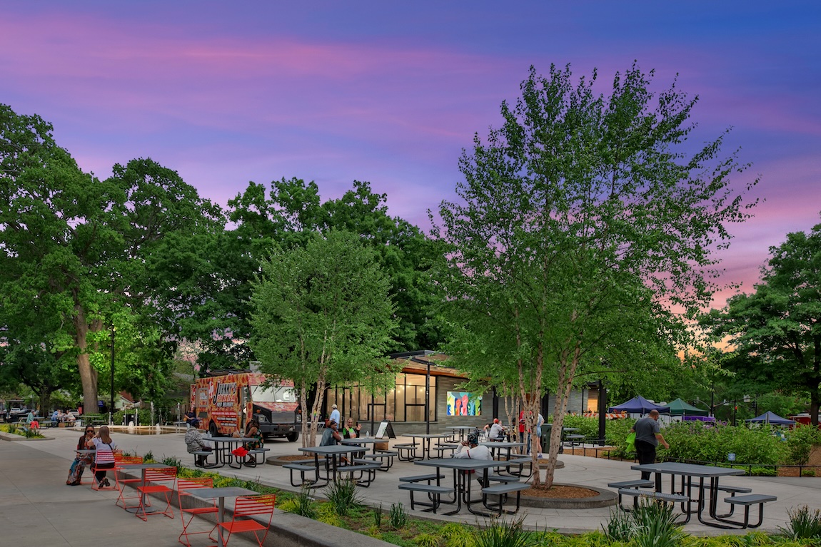 Community gathering space with outdoor seating, picnic tables, trees, and people enjoying a neighborhood plaza at dusk.