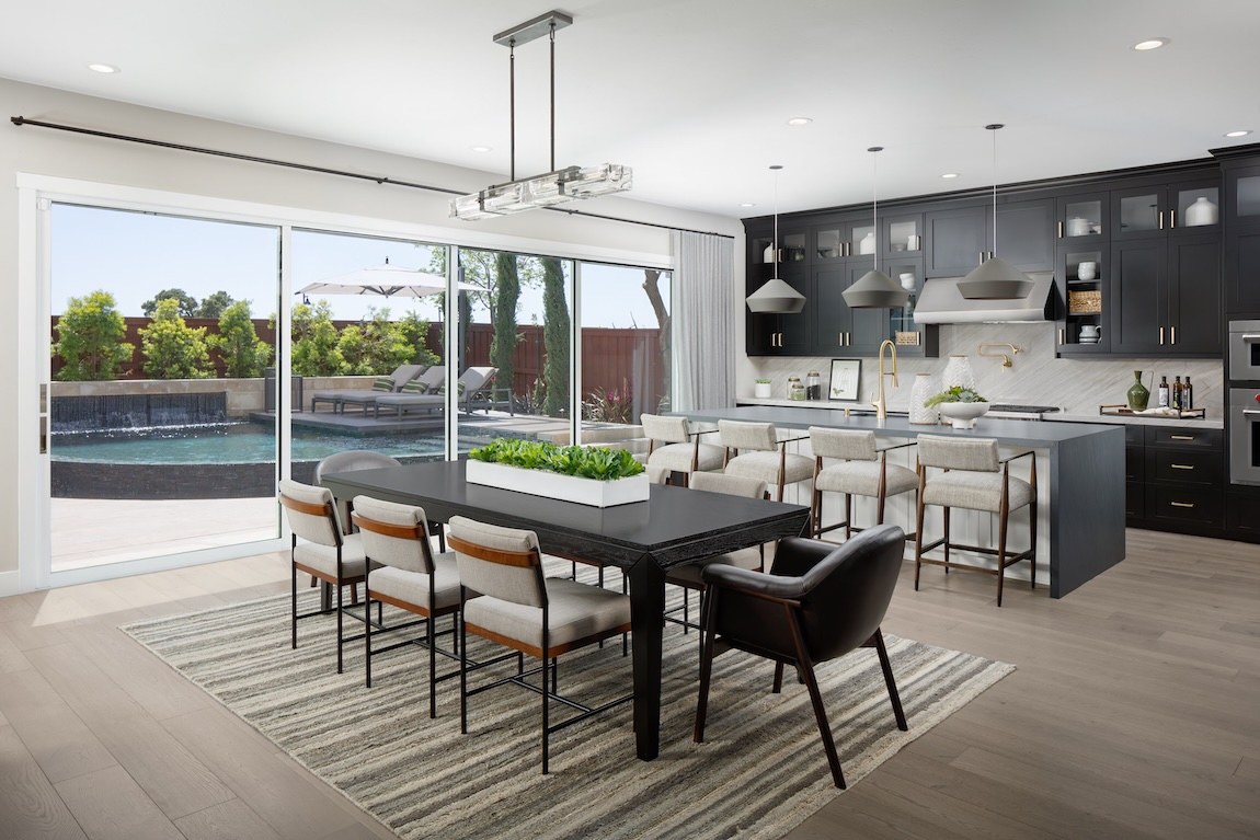 Bright open kitchen and dining space featuring a long island with upholstered bar stools, dark cabinetry, geometric pendant lighting, and expansive glass doors overlooking a backyard.