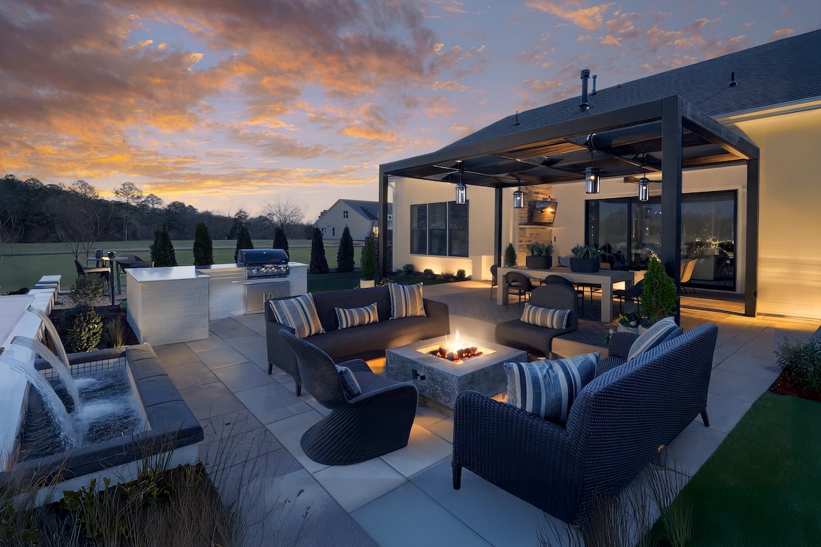Backyard outdoor living area with a covered patio, fire pit seating, outdoor kitchen, and landscaped yard during sunset.