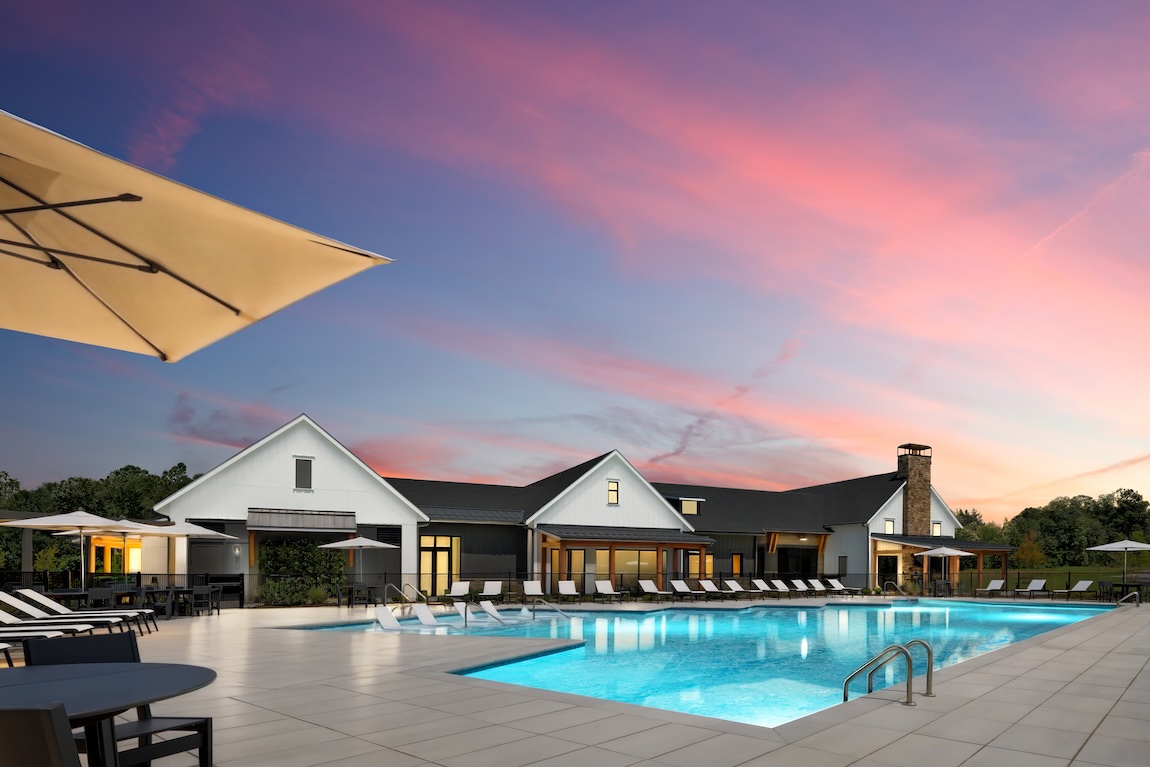 Resort-style community pool with lounge chairs, umbrellas, clubhouse buildings, and glowing water at twilight.