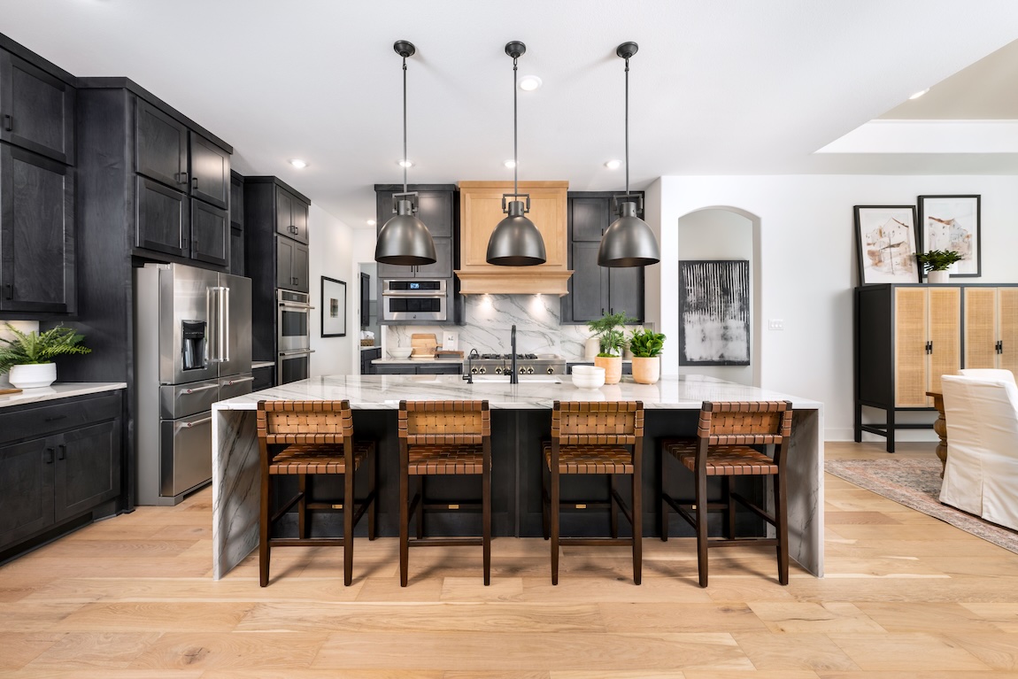 Modern kitchen with black cabinetry, marble backsplash, oversized island with woven leather bar stools, matte black pendant lights, and light hardwood floors.
