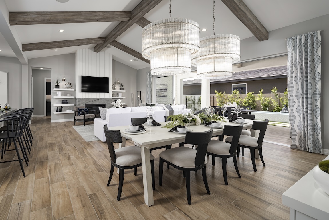 Open dining and living area with a long dining table, upholstered chairs, dramatic chandelier lighting, vaulted ceiling beams, and sliding doors opening to an outdoor space.