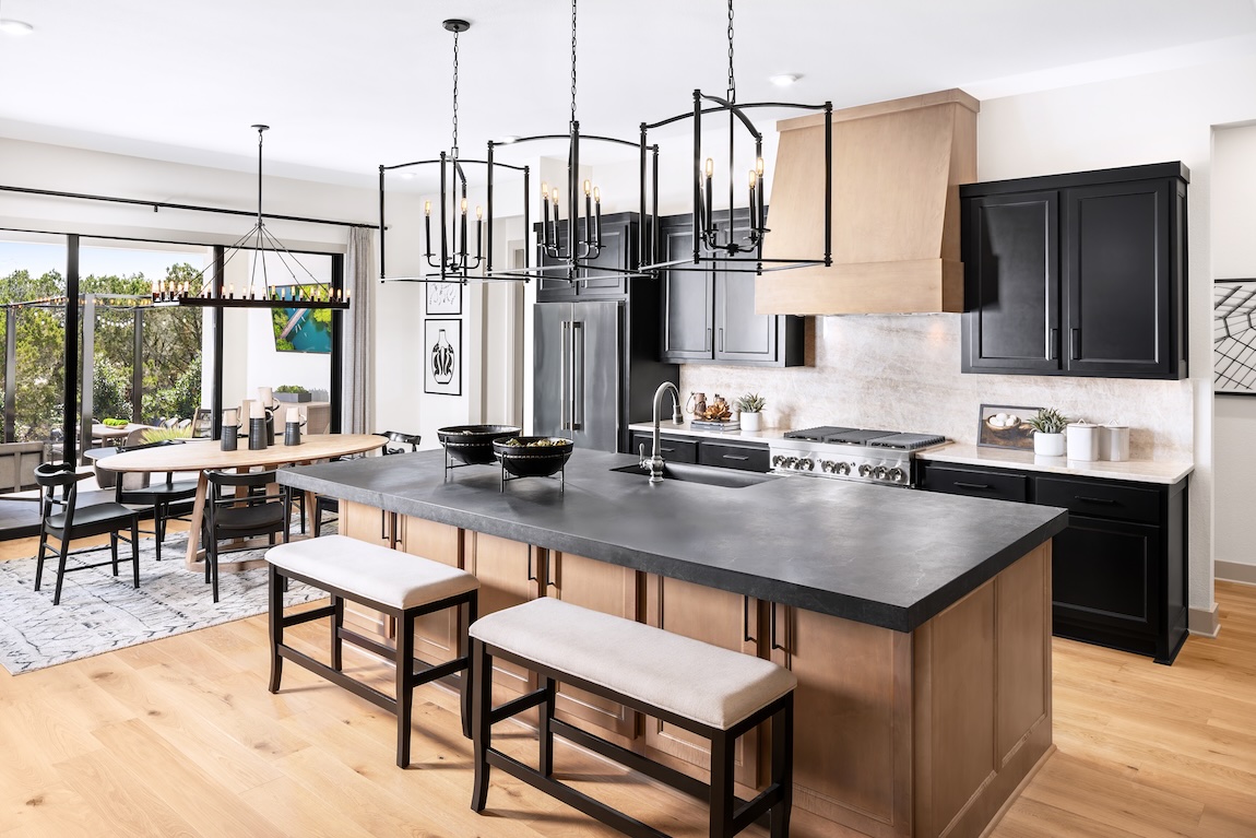 Contemporary kitchen with a charcoal island countertop, light wood base cabinets, black upper cabinetry, statement chandelier lighting, and an adjacent dining area.