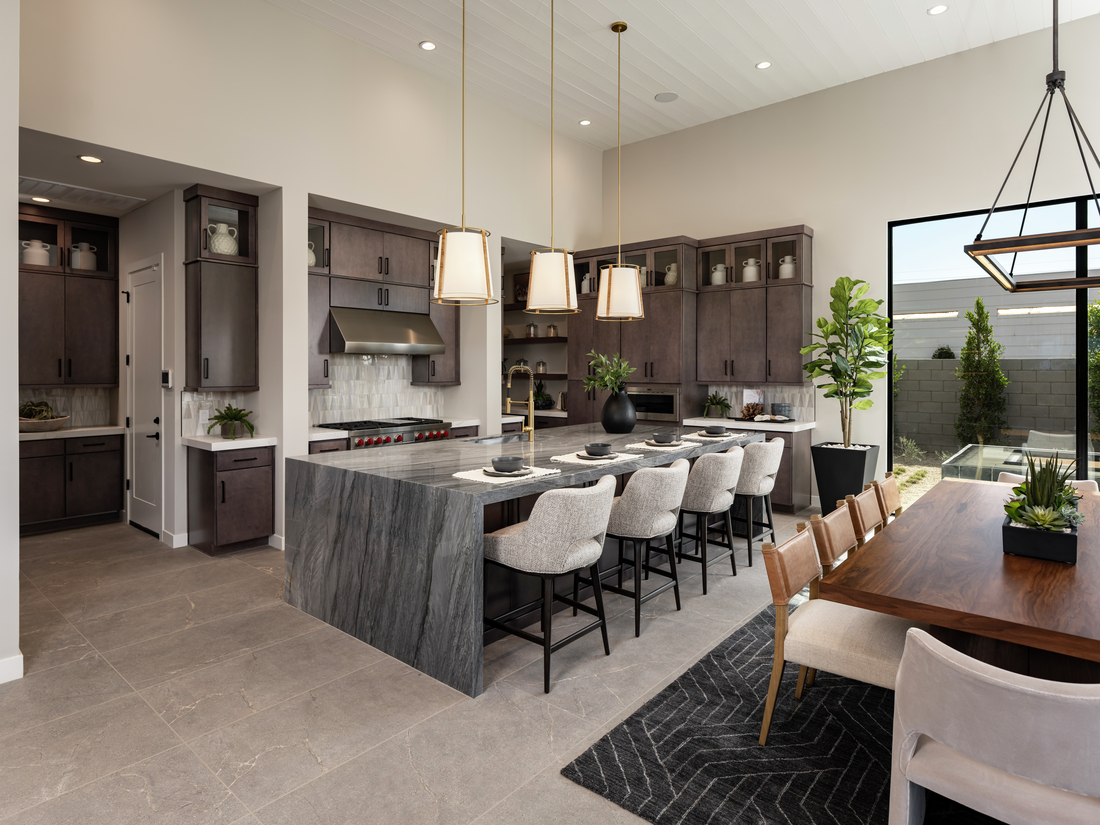 Open-concept kitchen and dining area with a dramatic gray stone island, soft gray cabinetry, sculptural pendant lights, and floor-to-ceiling windows bringing in natural light.