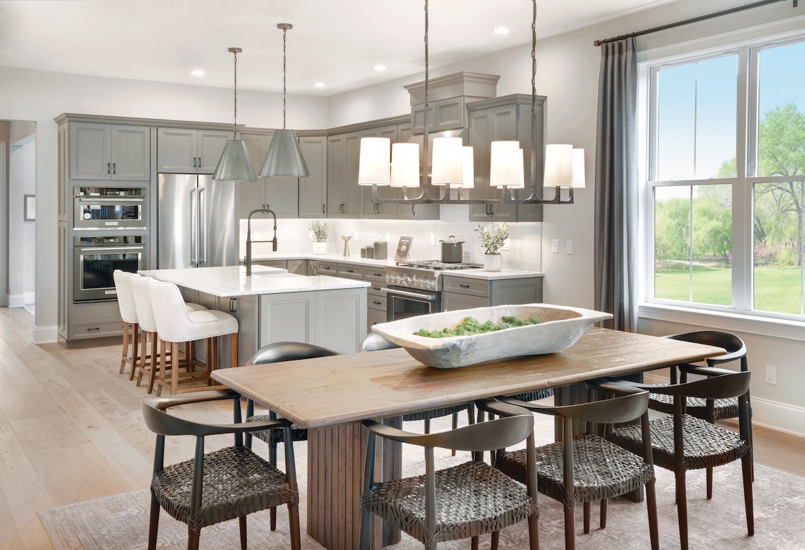 Open kitchen and dining area with soft gray cabinetry, white island with seating, modern pendant lighting, and a wood dining table near a large window.