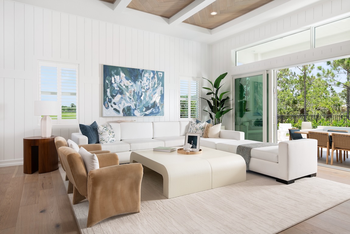 Bright living room with white paneled walls, a sectional sofa, light wood floors, vaulted ceiling details, and sliding glass doors to a patio.