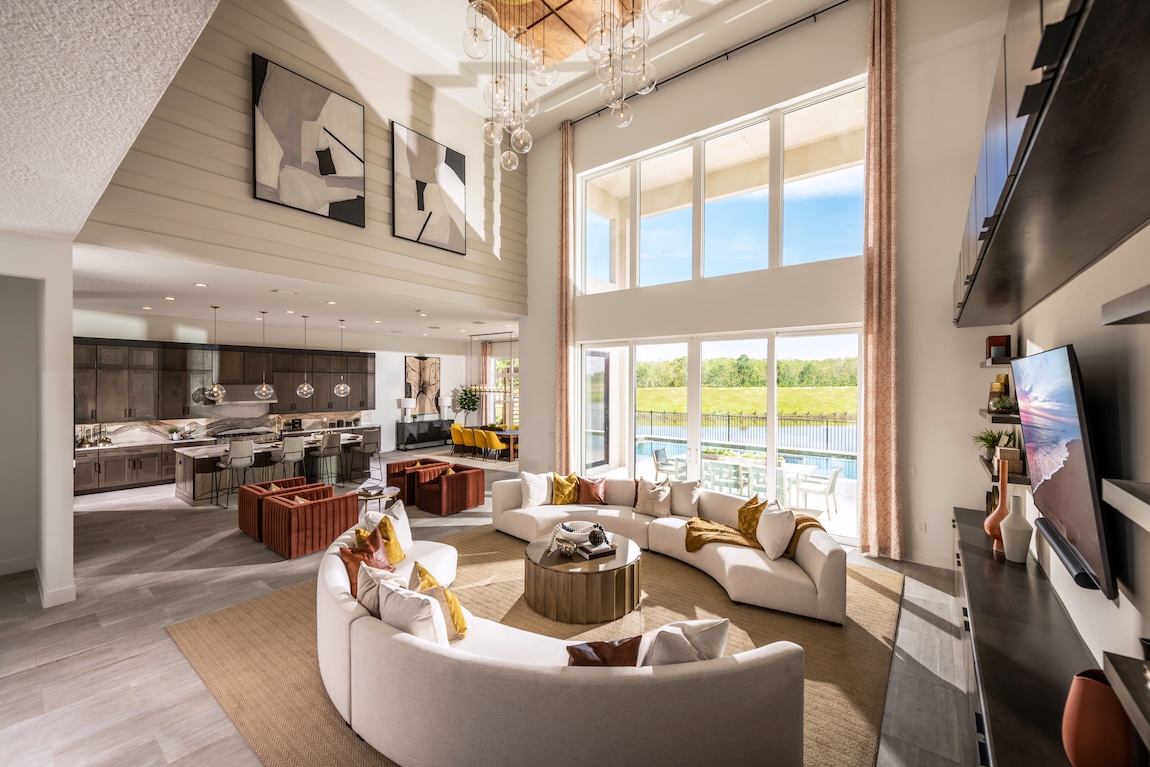 Expansive great room with curved sectional seating, double-height windows, modern chandelier lighting, and an open view into the kitchen and dining area.