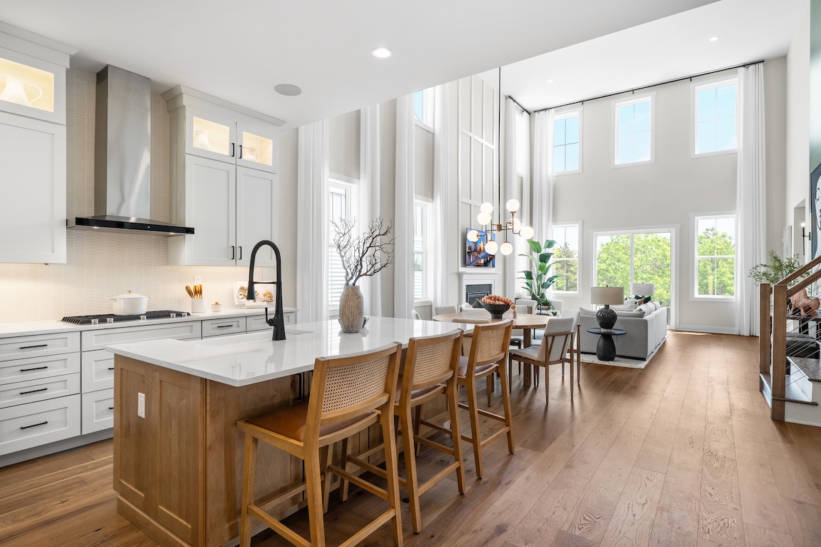 Open-concept kitchen and living area featuring a white island with wood base, cane-back bar stools, tall windows, and a cozy seating area beyond.