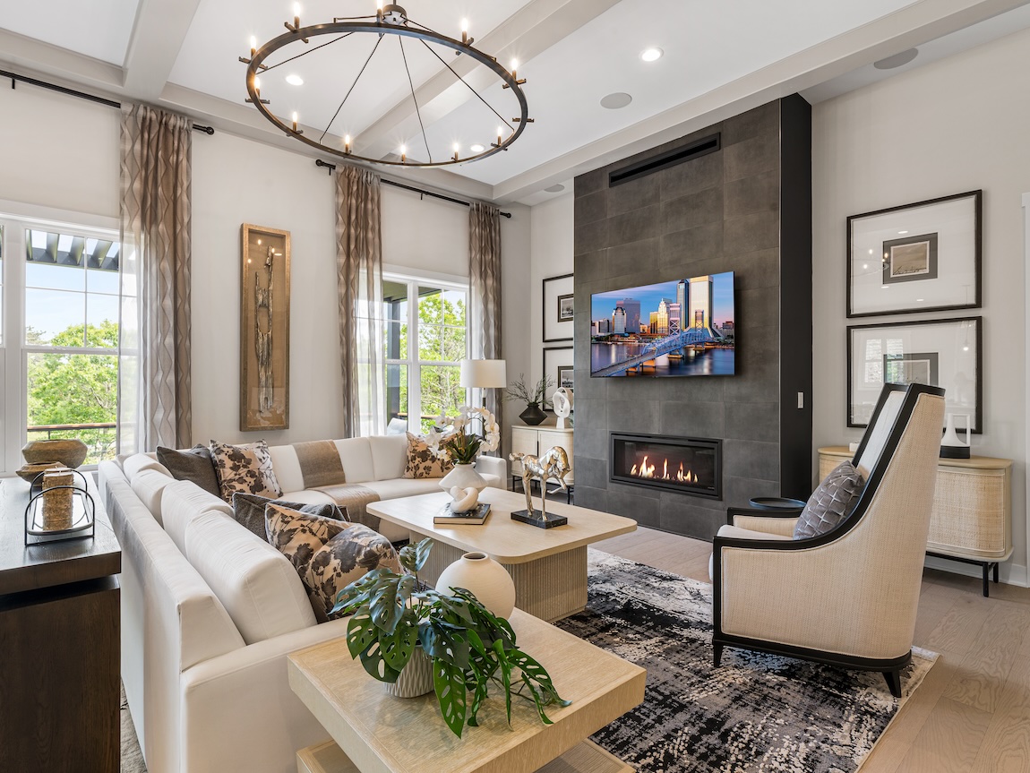 Stylish living room with a sectional sofa, modern fireplace wall, circular chandelier, layered neutral decor, and large windows overlooking greenery.