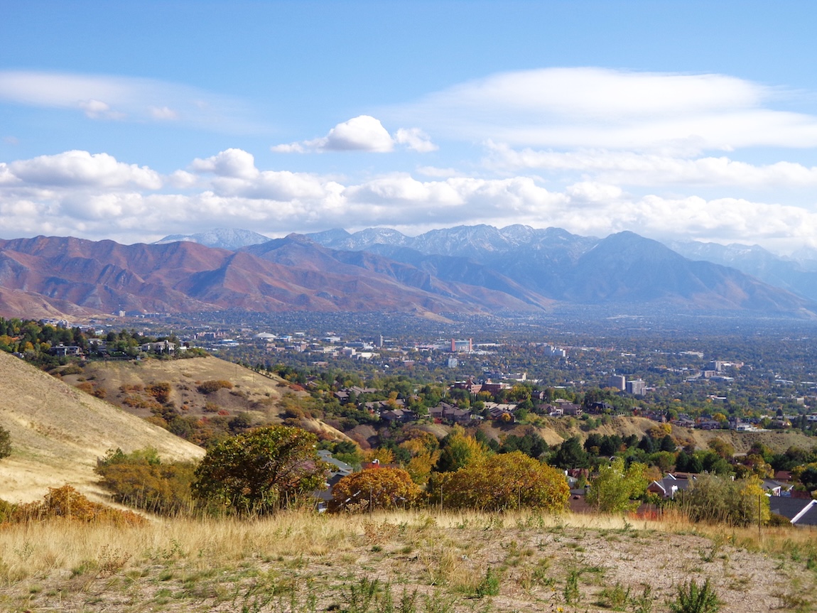 Panoramic mountain and valley view near Elk Ridge, Utah, featuring rolling foothills, autumn foliage, and snowcapped peaks beneath a bright blue sky.