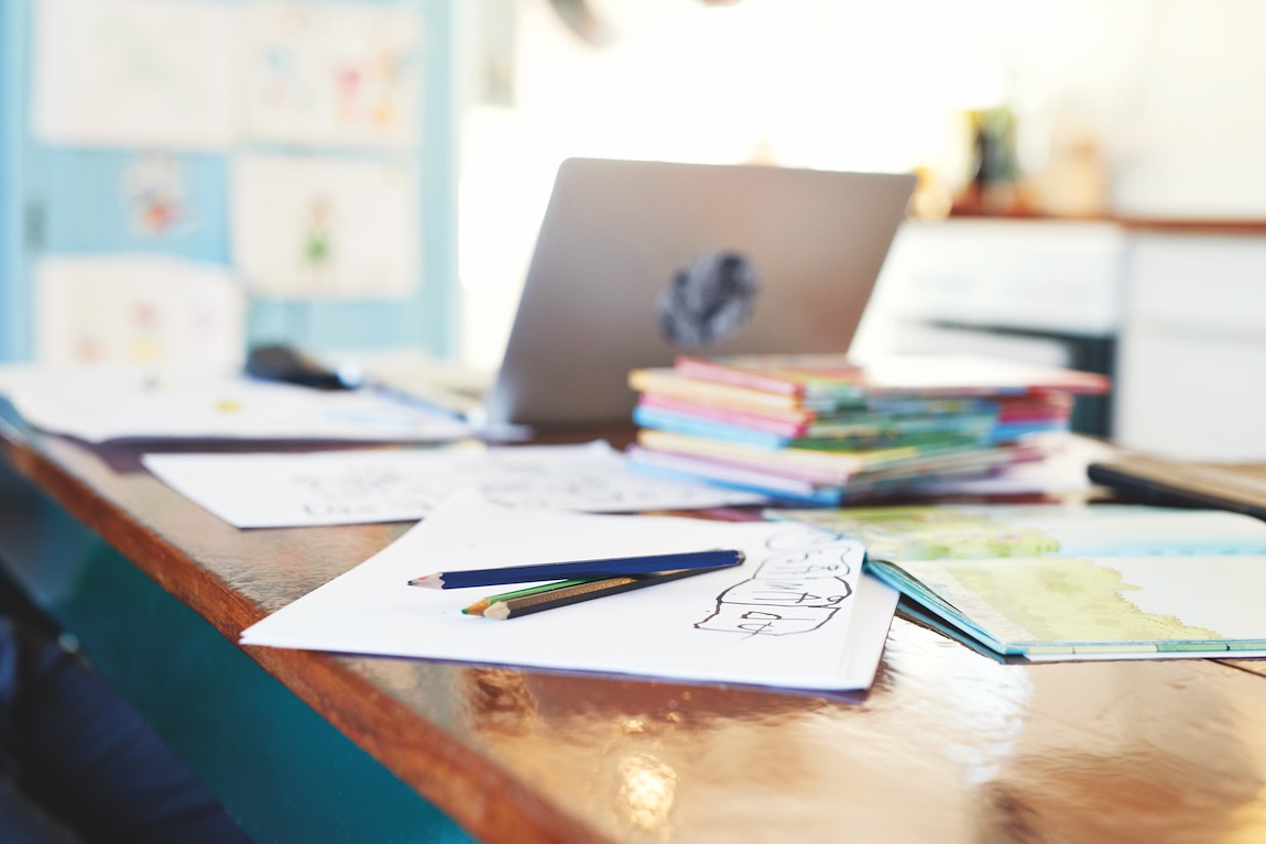 School workspace with children’s drawings, books, and laptop on a kitchen table.
