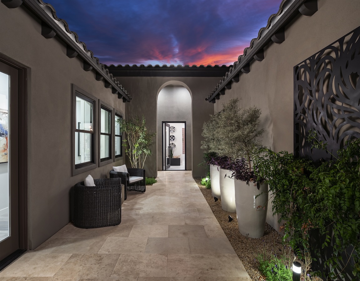 Tuscan-style courtyard entry with arched walkway, stucco walls, and lush landscaping at dusk.