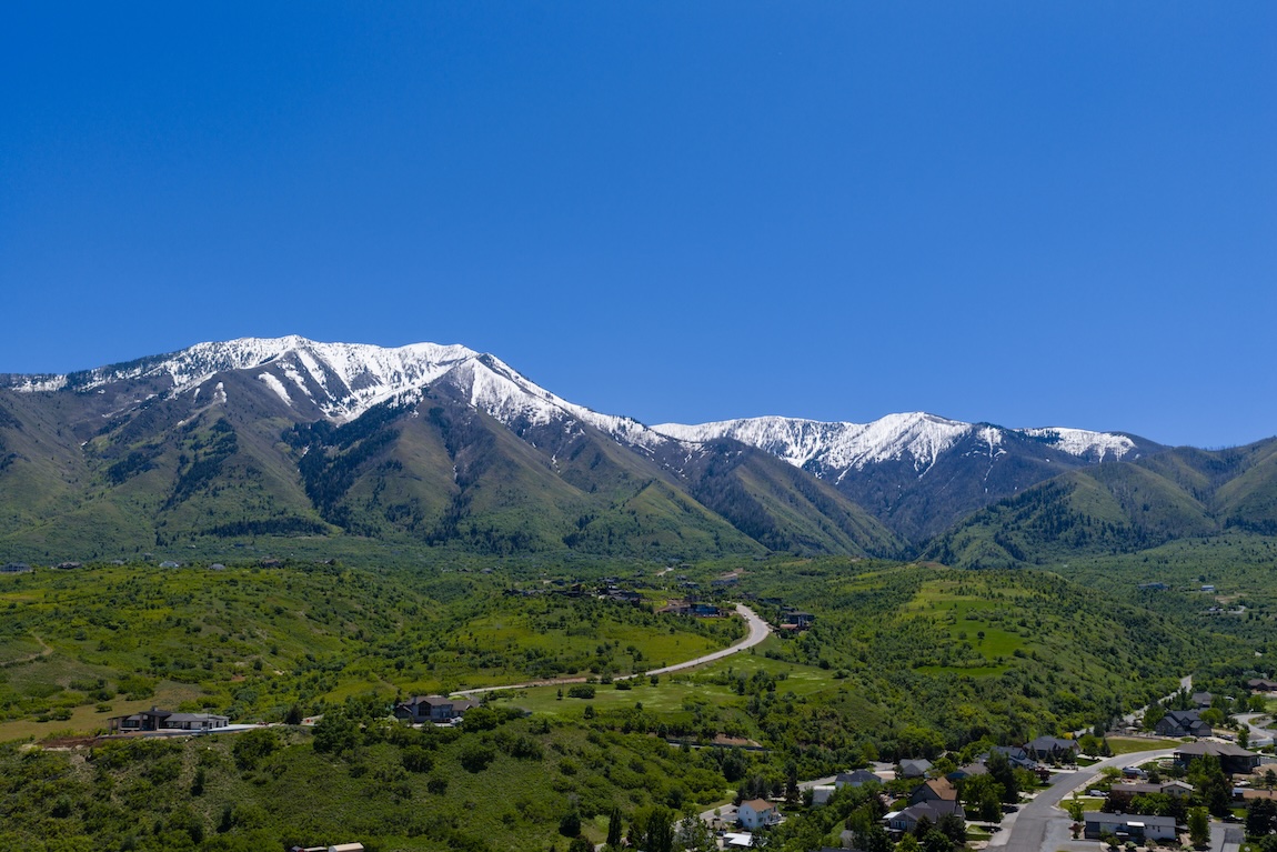 Mountain views surrounding Elk Ridge, Utah, with rolling green hills and snowcapped peaks under a clear blue sky.