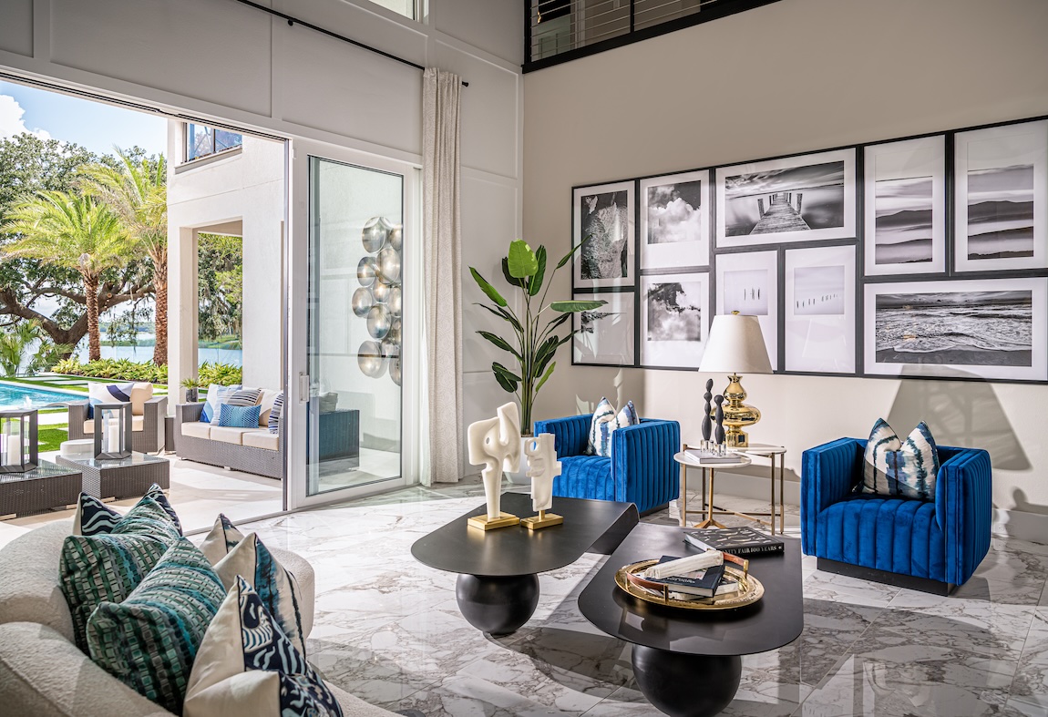 Double-height living room with vibrant blue accent chairs, marble flooring, bold artwork, and indoor-outdoor views.