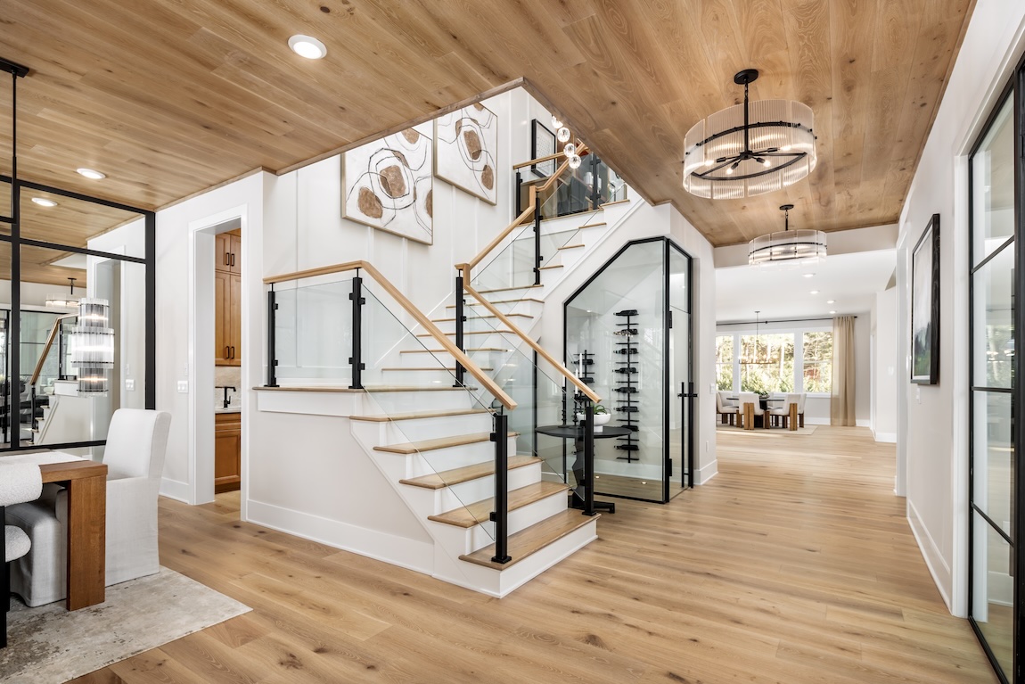 A light wood staircase with black metal and glass railings sits beneath a warm wood ceiling in an open-plan home.