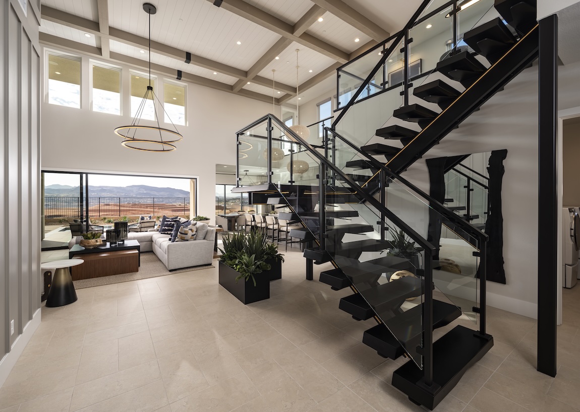 A striking black open-riser staircase with glass panels overlooks a bright living area and wide landscape views.