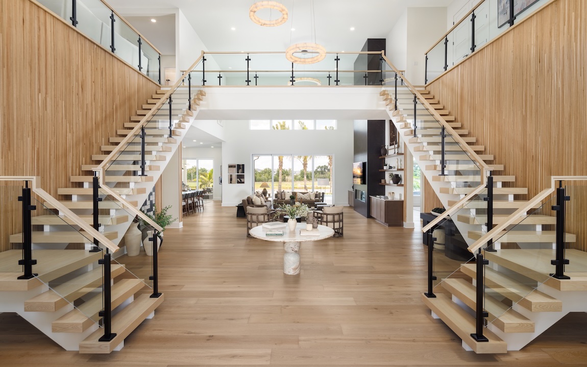 Two symmetrical staircases with light wood treads and dark railings frame a spacious, sunlit foyer.