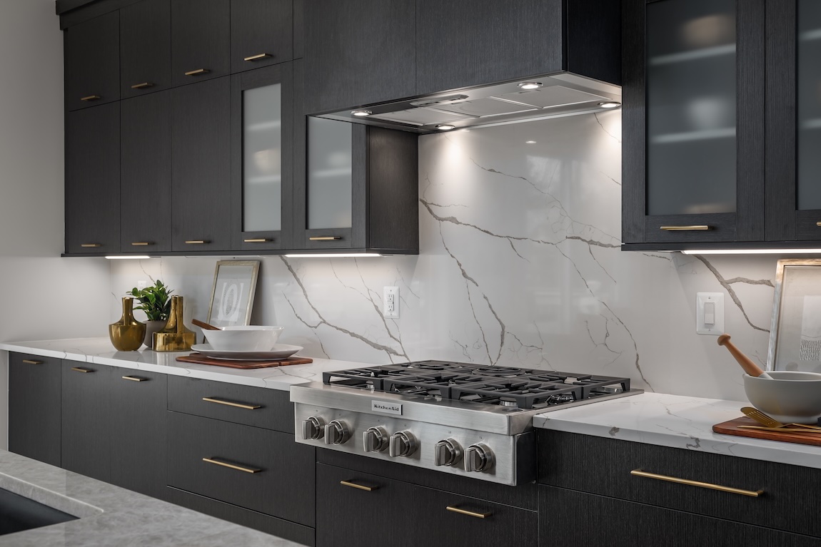 Sleek charcoal kitchen cabinetry with frosted glass inserts, brass hardware, and a dramatic marble-look backsplash surrounding a stainless steel cooktop.