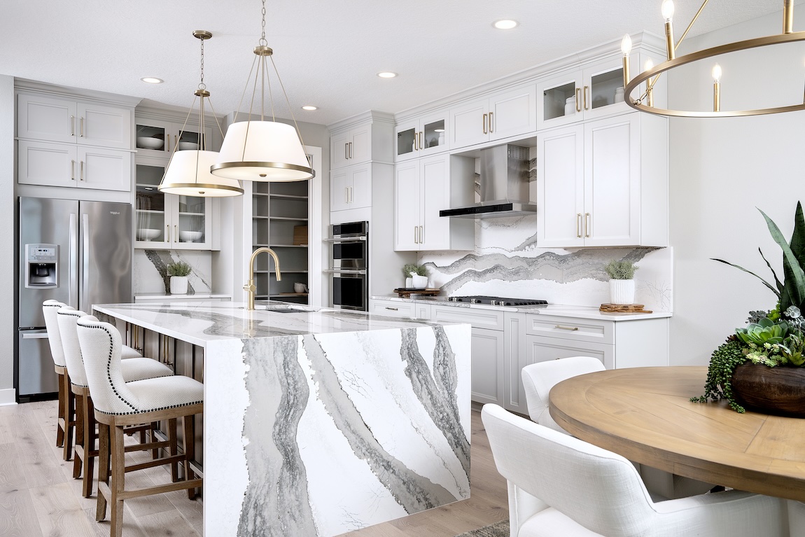Elegant white kitchen with glass-front upper cabinets, a statement stone backsplash, gold accents, and a waterfall island with bold gray veining.