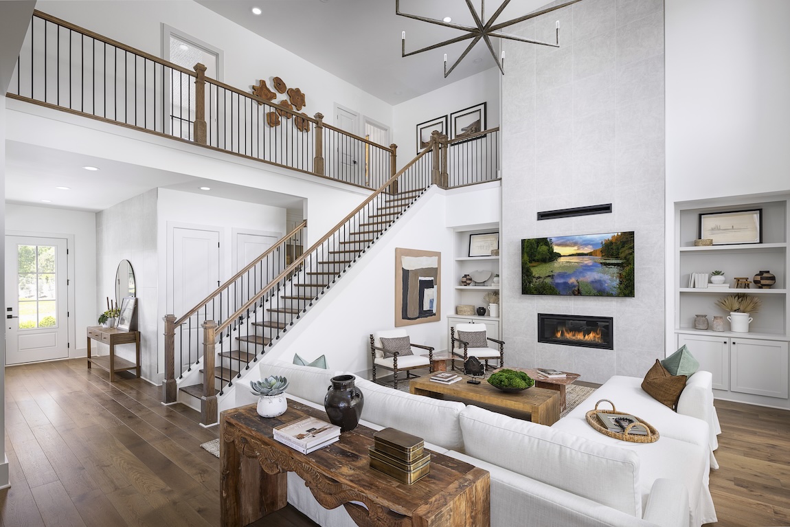A wood-and-metal staircase overlooks a two-story great room with a fireplace, white furnishings, and an upper-level gallery.