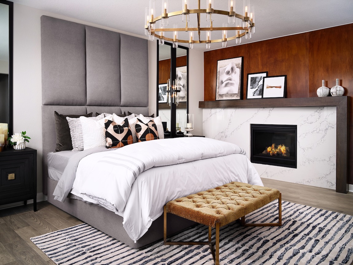 Luxury primary bedroom with an upholstered statement headboard, marble fireplace, brass chandelier, and warm wood accent wall.