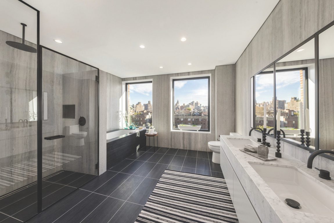 A modern bathroom with a bath, standing shower, daul-vanity display, and window with a view of New York City 