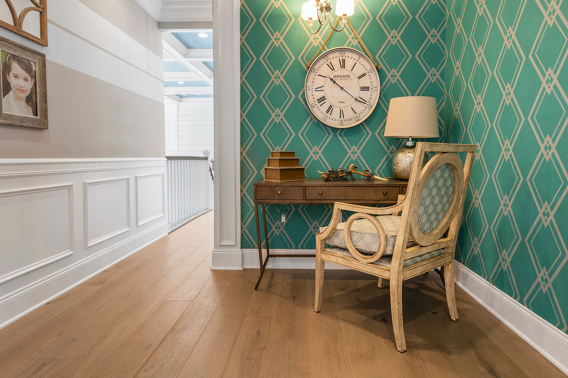 Writing desk in corner of hallway with green geometric wallpaper and large antique hanging clock.