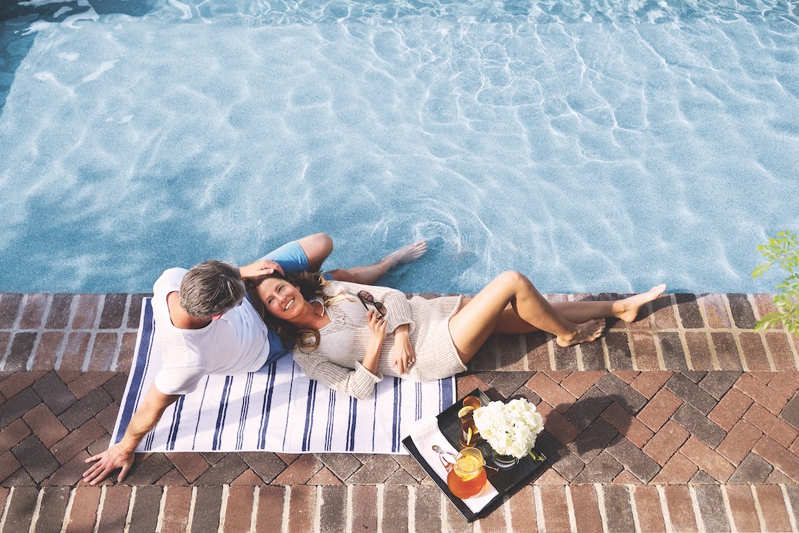 A man and woman sitting by the pool with a tray of iced tea and food