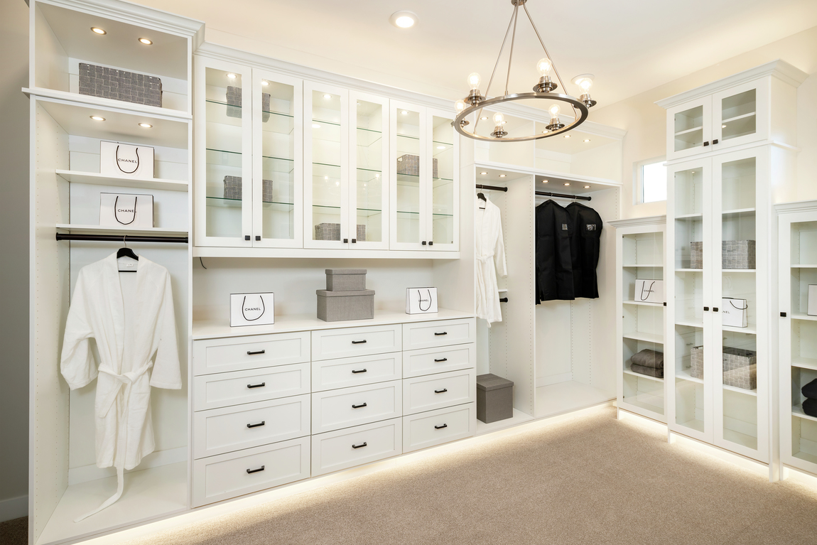 White closet cabinets with toe-kick lighting, spotlights, chandelier and glass cabinet doors. 