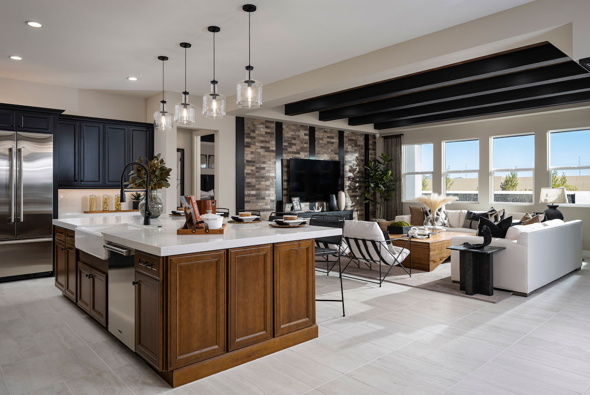 Open living space with kitchen and living room pictured. Kitchen island with four hanging pendant lighting fixtures and black accented wood beams. 